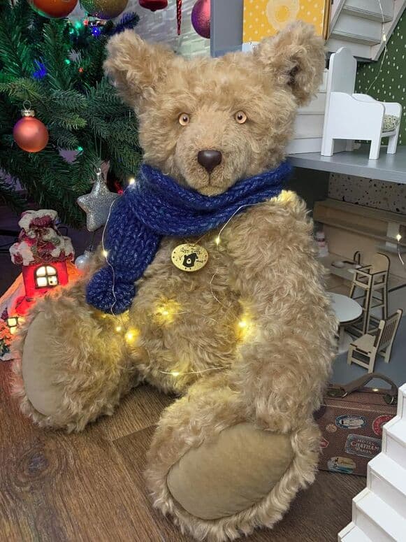 Noel, a sand-coloured mohair artist bear, seated by a Christmas tree